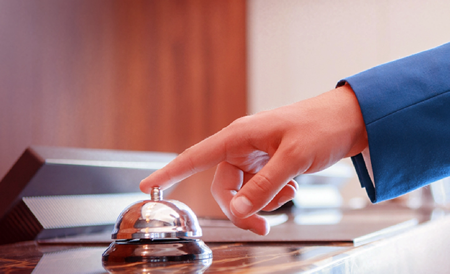 #BradescoAcessivel #PraTodoMundoVer: Uma mão de um homem que usa terno azul está com o dedo indicador sobre o botão da campanhia típica de balcões de hotel.