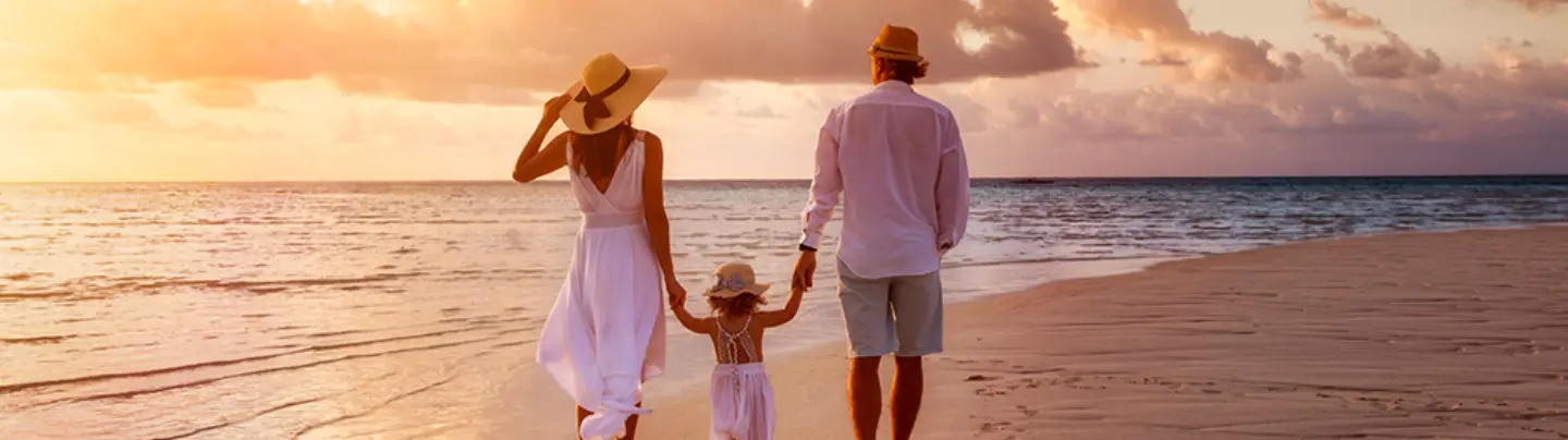 #BradescoAcessivel #PraTodoMundoVer: O pai e a mãe seguram as mãos da filha bem pequena enquanto andam na beira das ondas do mar descalços, de roupas brancas e de chapéu.