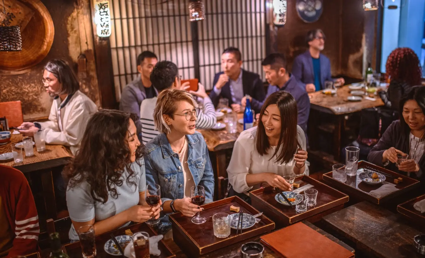 #BradescoAcessível #Pratodomundover Nas laterais, há grafismos vermelhos. No centro, três amigas estão em um bar japonês bebendo vinho e se divertindo.