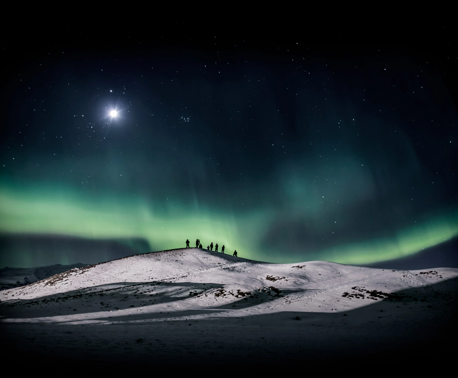 #BradescoAcessivel #ParaTodoMundoVer: Um grupo de pessoas em cima de um pequeno
    morro nevado forma a silhueta, ao observarem o céu com uma aurora bureal, presente
    nos polos do globo terrestre.