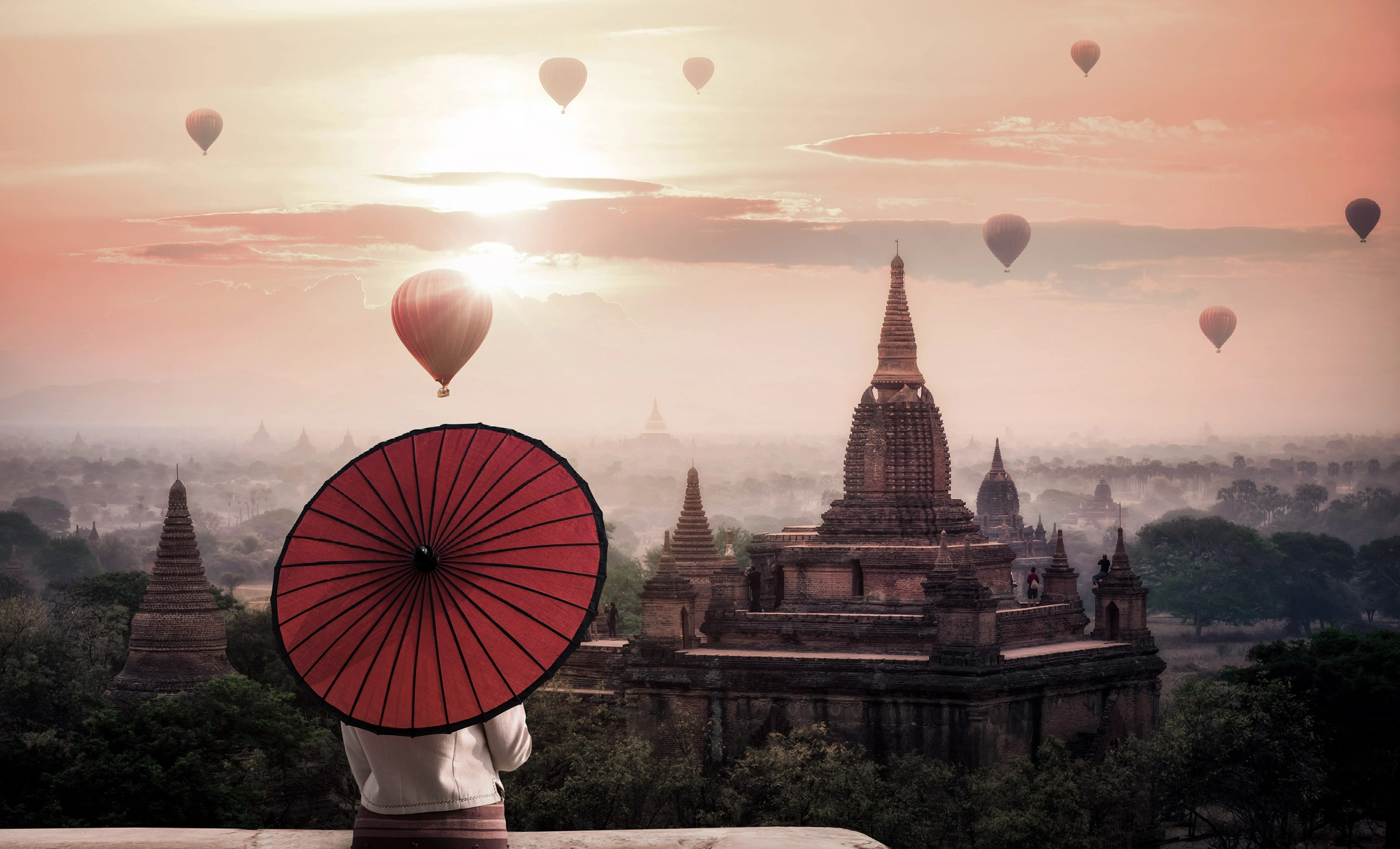 #BradescoAcessivel #ParaTodoMundoVer: uma pessoa segura um guarda-sol ao estilo
          japonês, cobrindo sua cabeça, enquanto olha vários balões que sobrevoam uma cidade
          asiática cheia de torres. Ao fundo, vemos um céu rosado com as nuvens cobrindo o
          sol.